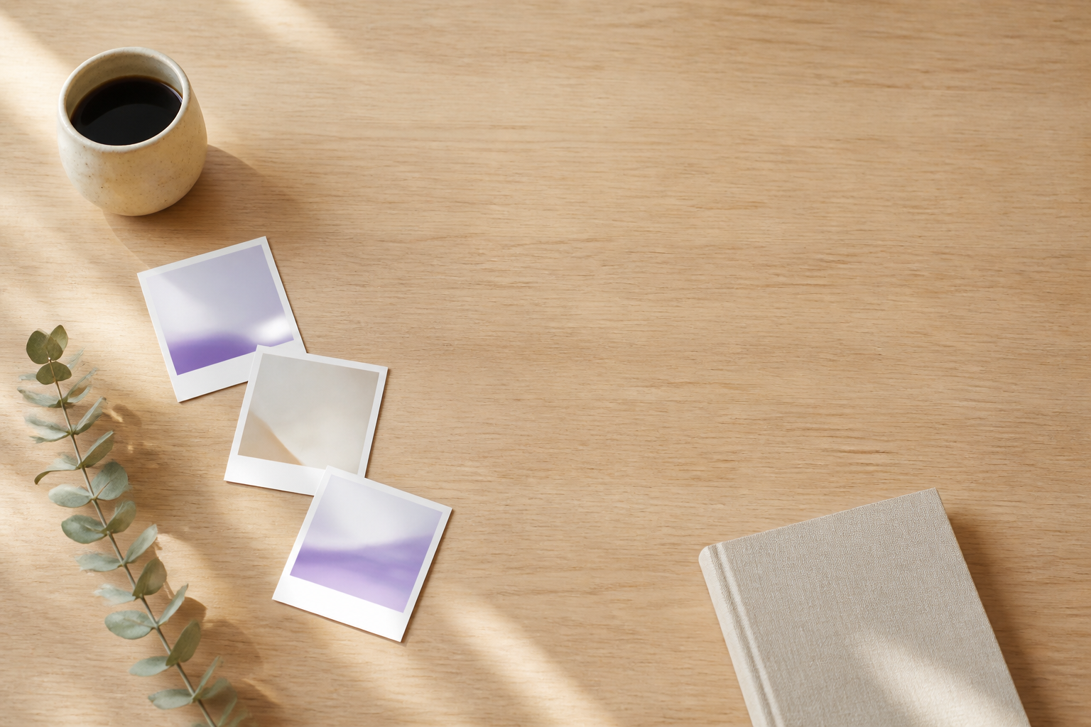 Abstract instant-photo prints, coffee, and a notebook on a sunlit oak table